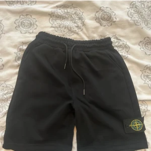 Svarta shorts från Stone Island - Svarta shorts från Stone Island med klassisk logopatch på benet. De har elastisk midja med snörning och två sidofickor. Materialet är mjukt och känns som bomull, perfekt för chill dagar. Snygg och enkel design som funkar till det mesta.