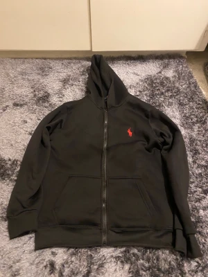 Svart kofta från ralph lauren - Märke: Polo Ralph Lauren Färg: Svart med rött broderat logo Storlek: mindre version av D Skick: Använd men i fint skick – inga hål eller fläckar Passform: Normal, unisex Detaljer: Dragkedja hela vägen, två stora fickor fram, mjukt och bekvämt material Perfekt för: Skolan, vardag, lager på lager, streetwear.                       hoodie 	•	zip hoodie 	•	svart hoodie 	•	svart ziptröja 	•	luvtröja 	•	basic hoodie 	•	streetwear 	•	herr hoodie 	•	unisex hoodie 	•	Polo hoodie 	•	sweatshirt 	