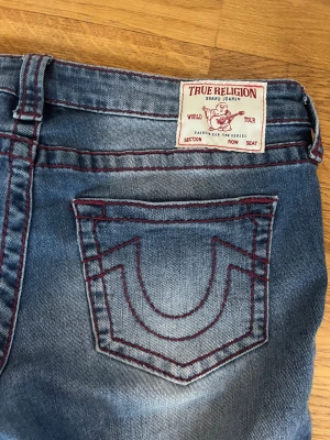 True Religion bootcut med röd sömn, stl 30 - Säljer ett par blå bootcut jeans från True Religion med ikoniska röda sömmar och tydliga bakfickor. Jeansen har klassisk femficksdesign, silverfärgade knappar och är tillverkade i slitstarkt denim. Perfekt för dig som gillar en avslappnad men snygg stil.