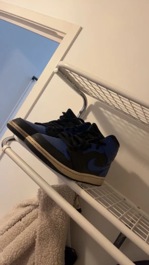 Nike Air Jordan 1 blå och svarta sneakers - Säljer ett par Nike Air Jordan 1 sneakers i blått och svart med klassisk swoosh på sidan. Skorna har snörning, rund tå och platt sula. Ovandelen är i skinn och de har en retro vibe som passar perfekt till streetwear. Ikonisk modell för dig som gillar sneakers med attityd.