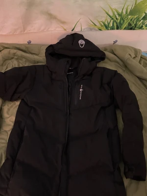 Svart dunjacka från Sail Racing - Svart dunjacka från Sail Racing med huva och dragkedja. Jackan är vind- och vattenavvisande, har andningsfunktion och är fodrad med certifierat dun (500 fill power). Snygg logga på bröstet och huvan, samt praktiska fickor med dragkedja. Perfekt för kalla dagar.