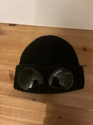Svart C.P. Company  mössa - Svart ribbstickad mössa från C.P. Company med ikoniska goggle-linser framtill. Mössan är tillverkad i mjuk ull och har en bred uppvikt kant. Perfekt för dig som vill sticka ut med streetwear-vibe och cool design.