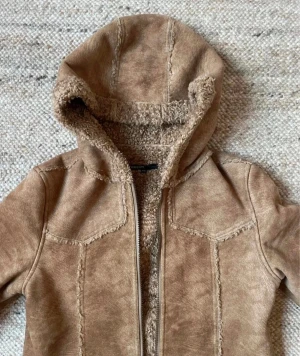 Beige kappa med huva från KappAhl - Säljer en beige teddyjacka från KappAhl med huva och mysigt fuskpälsfoder. Jackan har två stora fickor fram, synliga sömmar och dragkedja. Perfekt för dig som gillar chill och varm stil under kalla dagar.