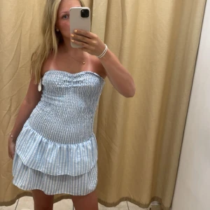 Sommarklänning - Säljer en söt axelbandslös kortklänning i blått och vitt med smockad överdel och volang i kjolen! Helt oanvänd!💞