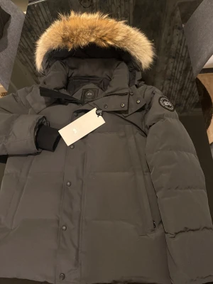 Canada goose Wyndham  - Säljer en svart dunjacka från Canada Goose med avtagbar huva och två olika pälskragar. Jackan har klassisk patch på ärmen, dragkedja och knappar framtill samt ribbade muddar. Perfekt för kalla vinterdagar och riktigt snygg streetstil. Väldig väldig varm och skön. Man får med båda pälsarna 
