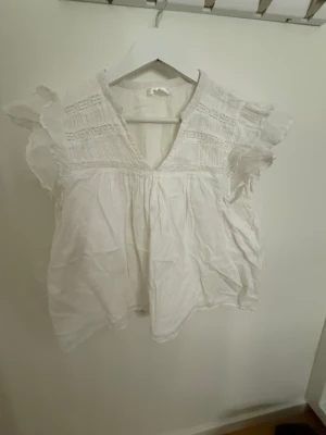 Vit blus med volangärmar och v-ringning - Säljer en söt vit blus med v-ringning och volangdetaljer på ärmarna, från Ginatricot. Blusen har broderade partier upptill och är tillverkad i ett lätt bomullstyg. Perfekt för sommarens alla tillfällen och ger en bra vibe till din outfit.