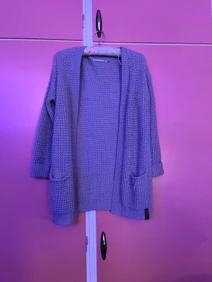 Ljuslila stickad kofta från 157 - Mysig gro stickad kofta från 157 med grovstickat mönster och två praktiska fickor framtill. Koftan har långärmade ärmar och ribbade muddar vid ärmslut och nederkant. Perfekt att slänga på sig när det är lite kyligt ute.