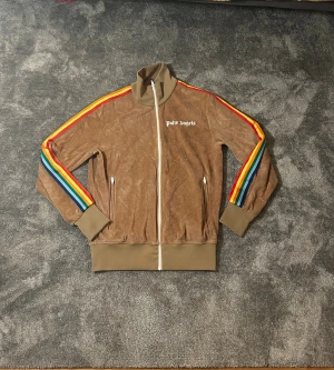 Palm Angels brun trackjacka S - Säljer en brun trackjacka från Palm Angels med coola regnbågsfärgade ränder längs ärmarna. Jackan har hög krage, vita dragkedjor och ribbade muddar.  Palm Angels-logga på bröstet. Liten slitning på sista bilden