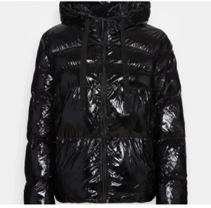 Svart vinterjacka från Guess - Säljer en svart, glansig pufferjacka från Guess med huva och snörning. Jackan har dragkedja framtill och är tillverkad i syntetmaterial. Guess-loggan syns på insidan. Perfekt för dig som gillar streetstyle och vill hålla dig varm. Pris kan diskuteras 