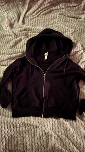 Svart hoodie med dragkedja - Svart hoodie med huva och snörning, hel dragkedja framtill och två fickor. Croppad passform med ribbade muddar vid ärmslut och nederkant. Perfekt för chill dagar och enkel att matcha med jeans eller mjukis.