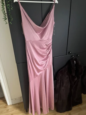 Glansig rosa balklänning - Säljer en glansig rosa balklänning med smala axelband och snygg drapering framtill. Klänningen har en cowl-neck ringning och är figurnära upptill med ett svepande fall nertill. Perfekt för dig som vill sticka ut med en elegant och trendig look.