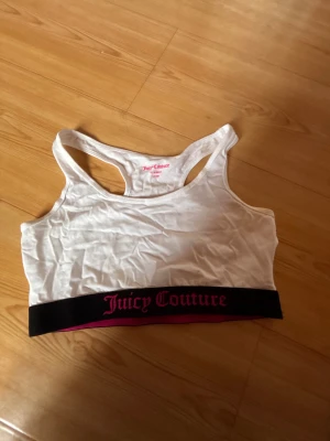 Vit linnetopp Juicy Couture 14-15 år - Säljer en vit linnetopp från Juicy Couture med bred svart resår och rosa logga längst ner. Toppen har sportig racerback och är tillverkad i mjuk bomull. Perfekt för dig som gillar streetstyle och vill ha något bekvämt och snyggt.