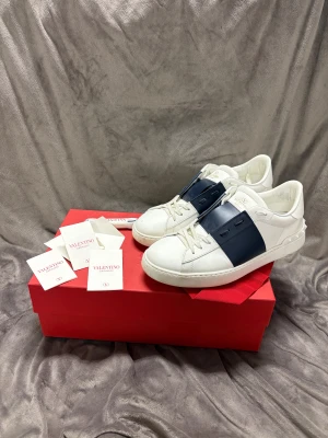 Valentino Garavani Open sneakers vit/blå - Snygga Valentino Garavani Open sneakers i vitt skinn med bred marinblå läderdetalj över vristen. Rund tå, platt sula och snörning framtill. Ikoniska nitar baktill på hälen ger extra edge. Perfekt för dig som gillar lyxiga och stilrena sneakers.