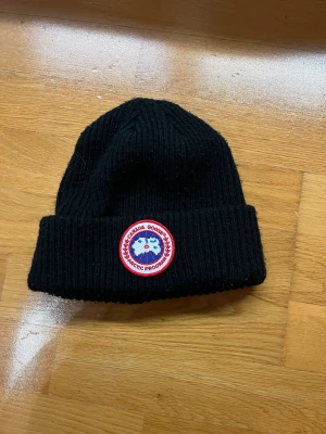 Svart ribbad mössa Canada Goose - Very good condition. Svart ribbstickad mössa från Canada Goose med klassisk rund patch framtill i rött, vitt och blått. Mössan har uppvikt kant och är tillverkad i mjukt stickat material som håller dig varm under kalla dagar. Perfekt för dig som vill ha en clean och stilren look.