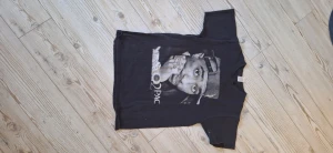 Svart 2Pac t-shirt med tryck - Svart t-shirt med stort 2Pac-tryck på framsidan. Klassisk passform med rund hals och korta ärmar. Perfekt för dig som gillar hiphop och streetwear. Snygg att matcha med jeans eller shorts.
