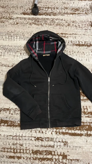 Svart Burberry kofta  - Svart hoodie från Burberry med klassiskt rutigt foder i huvan och broderad logga på bröstet. Tröjan har snörning i huvan, dragkedja framtill och två fickor. Materialet är mjuk bomull med fleece på insidan, perfekt för kyliga dagar. Säljer den för den är för liten för mig, den är i mycket bra skick och redo för sitt nya hem. Perfekt nu för vintern!