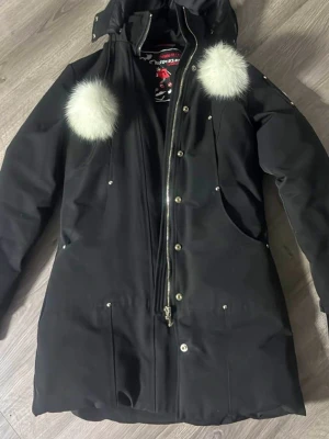 Svart dunjacka Moose knuckles - Svart dunjacka från Moose Knuckles med fluffiga vita pälsdetaljer på huvan och axlarna. Jackan har dragkedja, knappar och ett metallmärke på ärmen. Insidan är quiltad och fodrad för extra värme. Perfekt för kalla dagar och har en snygg, lång siluett.  Kan tänkas sänkas vid snabb affär.