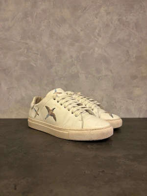 Axel Arigato sneakers - Säljer ett par vita sneakers från Axel Arigato med Beebird design på sidorna och ovansidan. Skorna har rund tå, platt sula och snörning. Tillverkade i skinn med en stilren och minimalistisk design, perfekt för dig som gillar detaljer.