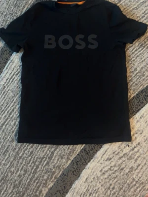 Svart BOSS t-shirt med tryck - Svart t-shirt från BOSS med stort, ton-i-ton tryck över bröstet. Klassisk rund hals och korta ärmar. Materialet känns mjukt och skönt, perfekt för en chill look. Snygg och enkel design som funkar till jeans eller shorts.