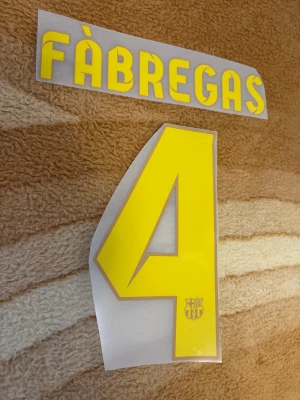 Barcelona Fàbregas 4 tryck - Säljer ett officiellt Barcelona-tryck med FÀBREGAS och nummer 4 i gul färg med grå kant. Perfekt för att stryka på din matchtröja. Numret har en liten Barca-logga längst ner.