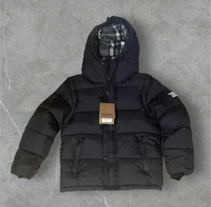 Svart pufferjacka från Burberry - Svart pufferjacka från Burberry med huva och klassiskt rutigt foder. Jackan har dragkedja och knappar framtill, två fickor med dragkedja och ribbade muddar vid ärmslut. Perfekt för kalla dagar och har en snygg, modern look. Pris kan diskuteras 