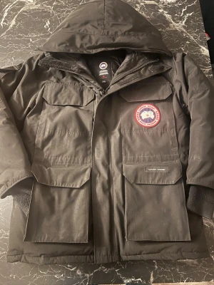 Svart Canada Goose Expedition dunjacka - Svart Canada Goose Expedition dunjacka med huva, stora fickor framtill och klassisk röd logotyp på bröstet. Jackan är fodrad med dun och fjädrar, har dragkedja och kardborre framtill samt ribbade muddar vid ärmslut. Perfekt för kalla vinterdagar. INGA RETUR ACCEPTERAS