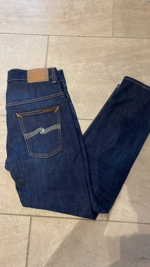 Nudie Jeans Thin Finn blå jeans - Säljer ett par Nudie Jeans Thin Finn i mörkblå tvätt med klassisk orange söm och broderad bakficka. Modellen är slim och har en rak passform med normal midja. Materialet är 98% bomull och 2% elastan för extra stretch och komfort. 33/30