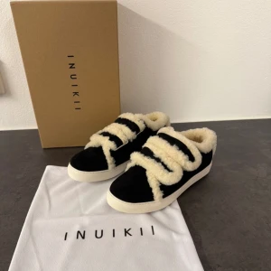 Svarta Inuikii sneakers med fårskinn - Unika svarta sneakers från Inuikii med fluffigt beige fårskinn både på insidan och utsidan. Skorna har breda kardborreband täckta av fårskinn och en vit platt sula. Perfekta för dig som vill ha något riktigt cozy och trendigt till höst och vinter.