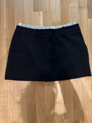 Svarta skorts från Zara med randig midja - Snygga svarta skorts från Zara i storlek L. De har en omlott-design framtill och en elastisk midja med vitt tyg och svarta ränder. Perfekt för dig som vill ha shorts med kjol-look. Materialet är mjukt och bekvämt, och midjan har hällor för bälte.
