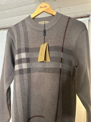 Beige rutig stickad tröja Burberry - Snygg stickad tröja från Burberry i beige med klassiskt rutigt mönster i grått och brunt. Rund halsringning och långa ärmar. Perfekt för dig som gillar stilrena designer och vill ha något som sticker ut lite extra.