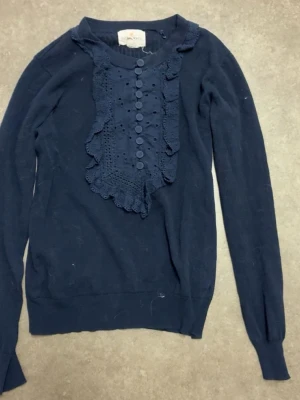 Mörkblå stickad tröja Holly & Whyte - Supersnygg mörkblå stickad tröja från Holly & Whyte med lång ärm. Framsidan har dekorativa knappar och spetsdetaljer i samma färg, vilket ger en cool och unik look. Perfekt för dig som gillar detaljer och vill ha något extra i garderoben.