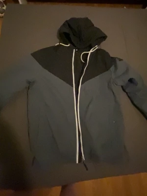 Svart och mörkgrå pufferjacka FSBN L - Snygg pufferjacka från FSBN i storlek L, med svart och mörkgrå färgblock, huva med snörning och vit dragkedja