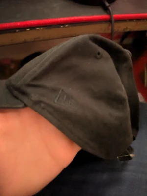 Svart keps från New Era - Säljer en stilren svart keps från New Era med justerbart spänne baktill. Kepsen har en klassisk form och är tillverkad i bomullsmaterial. Perfekt för dig som vill ha en enkel och snygg accessoar till din outfit.