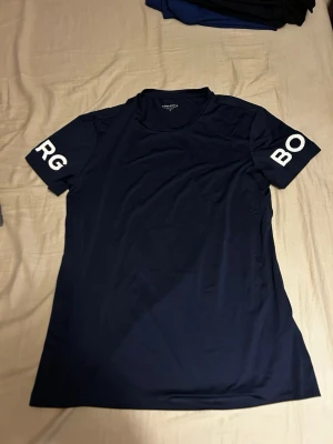 Mörkblå t-shirt från Björn Borg - Snygg mörkblå t-shirt från Björn Borg med vit logotyptext på båda ärmarna. T-shirten har rund hals och är tillverkad i mjukt polyester-material som andas, perfekt för träning eller chill. Enkel och stilren design som funkar till det mesta.