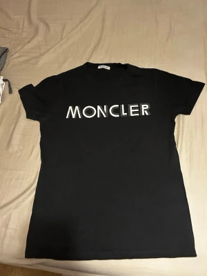 Svart Moncler t-shirt med tryck - Svart t-shirt från Moncler med stort vitt logotryck över bröstet. Klassisk rund hals och korta ärmar. T-shirten har en clean och stilren look som passar perfekt till jeans eller shorts. Materialet känns mjukt och bekvämt mot huden.