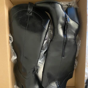 Svarta cowboy boots med klack - Säljer ett par svarta cowboy boots med spetsig tå och hög klack. Skorna har använts en gång och är i ett mycket bra skick. Finns en liten repa som man kan se på bild fyra men annars är de som nya. Nu pris 780 kr. 