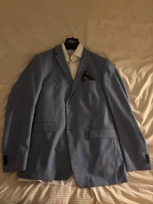Tiger of Sweden kostym  - Elegant kostym i ljusblå färg. Kavajen har en klassisk passform med knäppning framtill och fickor. Tillhörande byxor i matchande färg ingår. En vit skjorta från Eton i slim fit storlek 41/16 och en orange slips ingår också. (Tiger of Sweden) OBS! Det är ingen röd fläck på kostymen på första bilden, då kameran är så! 