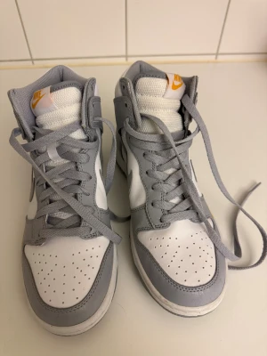 Nike Dunk High vit och grå sneaker - Nike Dunk High sneakers i vitt och ljusgrått med högt skaft och klassisk snörning. Skorna har en rund tå, perforerade detaljer på tån och orange Nike-logga på plösen och sidan. Tillverkade i skinn och syntetmaterial för en clean och sportig look.