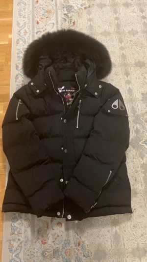 Svart Moose Knuckles dunjacka med päls - Svart dunjacka från Moose Knuckles med fluffig päls på huvan, silverfärgade dragkedjor och logga på ärmen. Jackan har flera fickor med dragkedja och snygga detaljer som tryckknappar och broderad insida. Perfekt för kalla dagar och riktigt stilren look. För erat info jag är 181 cm lång jag väger 63 kg och passar perfekt 