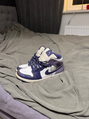 Nike Air Jordan 1 Mid vit och lila - Nike Air Jordan 1 Mid sneakers i vit och mörkblå färg med klassisk snörning och rund tå. Skorna har en ikonisk Jordan-logga på sidan och är tillverkade i skinn med perforerad tå för extra ventilation. Perfekt för dig som gillar streetwear och vill sticka ut. Passar även 45