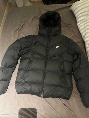 Svart Nike pufferjacka Storm-FIT M - Svart pufferjacka från Nike med Storm-FIT teknologi. Jackan har huva, två sidofickor med dragkedja och en klassisk Nike-logga på bröstet. Perfekt för kalla dagar och har en schysst, lite loose passform. Materialet är polyester och jackan stängs med dragkedja.