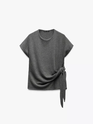 Mörkgrå topp med knut från Zara - Säljer en stilren mörkgrå topp från Zara i storlek M. Toppen har korta ärmar, rund hals och en snygg knytning i sidan som ger en cool detalj. Materialet är mjukt och stickat, perfekt för dig som gillar enkel men trendig design.