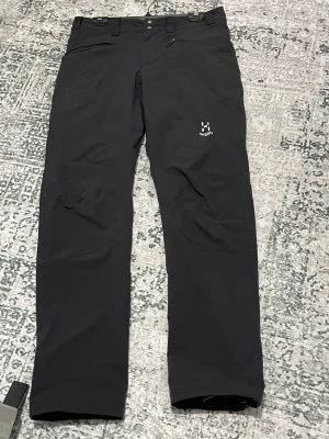  Haglöfs byxor  - Svarta täckbyxor från Haglöfs med raka ben och flera praktiska dragkedjor på fickorna. Byxorna har normal passform och är tillverkade i slitstarkt syntetmaterial, perfekt för utomhusaktiviteter. Snygg logga på låret och bälteshällor i midjan.