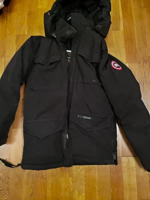 Svart Canada Goose parkajacka - Svart parkajacka från Canada Goose med ikonisk patch på ärmen. Jackan har flera stora fickor framtill, huva och dragkedja. Perfekt för kalla vinterdagar och har en clean, klassisk look som funkar till streetwear. Materialet är slitstark polyester och dunfyllning.