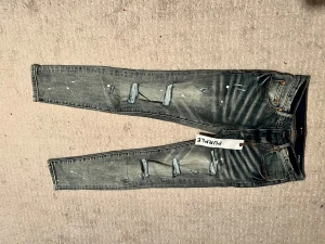 Blå skinny jeans från Purple Brand - Säljer ett par blå skinny jeans från Purple Brand med slitna detaljer och färgstänk på både fram- och baksida. Jeansen har klassisk femficksdesign, låg midja och dragkedja. Materialet är jeans med en tvättad look och råa hål på benen för en edgy stil.