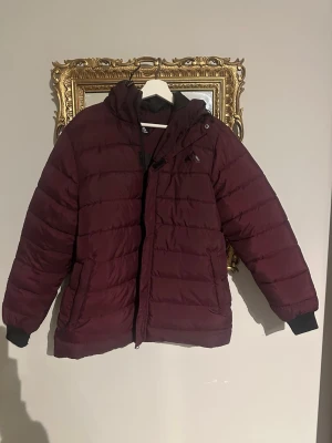 Vinröd pufferjacka från High Mountain - Säljer en vinröd pufferjacka från High Mountain i damstorlek 42. Jackan har huva, svarta muddar vid ärmslut och dragkedja framtill. Materialet är syntet och jackan är quiltad för extra värme. Perfekt för kalla dagar och har en clean look.