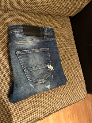 Blåa jeansshorts från Hoodrich - Säljer ett par blåa jeansshorts från Hoodrich med slitna detaljer och broderad HR-logga på bakfickan. Shortsen har klassisk femficksdesign, knappgylf och text på insidan av linningen. Perfekta för en avslappnad streetstil.