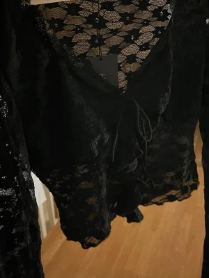 Svart spetsblus  - Säljer en svart blus med transparent spets och snyggt knyt framtill. Blusen har lång ärm och v-ringad hals, samt ett croppat snitt som ger en cool vibe. Perfekt för dig som gillar detaljer och vill sticka ut lite extra.