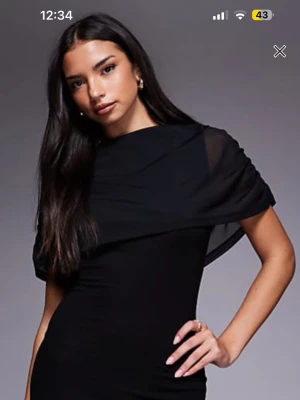 Svart långklänning med draperad mesh - Snygg svart långklänning med draperad mesh över axlarna och övre delen av ryggen. Klänningen har en figurnära passform och är tillverkad i ett mjukt, stretchigt material. Meshdetaljen ger en cool och elegant look, perfekt för dig som gillar stilrena plagg med en twist. Aldrig använd