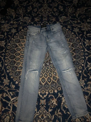 Supply & Demand ljusa ripped jeans XS - Snygga ljusblå jeans från Supply & Demand med slitna detaljer och ripped effekt på benen. Modellen har klassisk femficksdesign, knapp och dragkedja fram. Perfekt för dig som gillar streetstyle och vill ha en cool look.